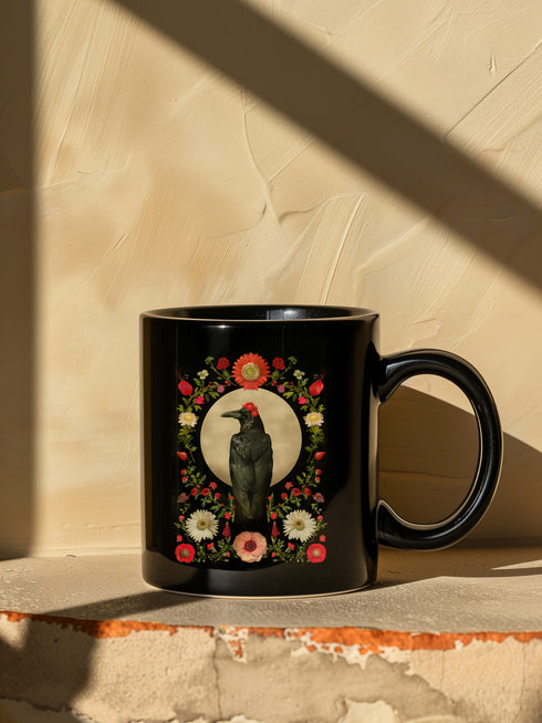A black ceramic mug featuring a gothic design of a raven against a golden full moon, framed by a floral wreath of coral-colored daisies, white chrysanthemums, and delicate botanical elements. The design includes rich green foliage and small red buds arranged in a symmetrical pattern. The mug has a classic C-shaped handle with a glossy finish.